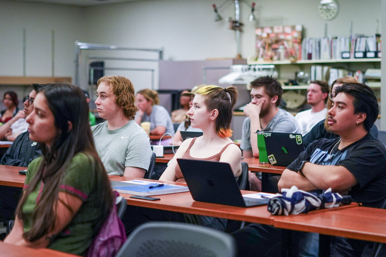 A group of young adults are seated at desks in a classroom setting, attentively listening or looking forward. Several students have laptops on their desks.